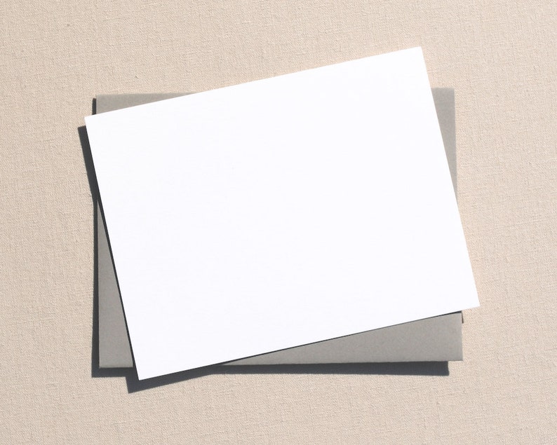 May include: A blank white card with rounded corners is placed on top of a gray envelope. The envelope and card are on a beige textured surface.
