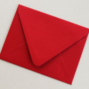 May include: A vibrant red envelope with a triangular flap, partially open. The envelope is made of smooth paper and rests on a light beige textured surface. The image presents a clean, simple composition, suitable for stationery or correspondence.