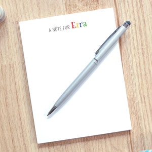 May include: A white notepad with the text "A note for Ezra" in colorful letters. A silver pen with a stylus tip is on top of the notepad.