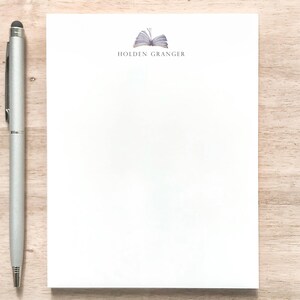 May include: A white notepad with a book icon and the text "HOLDEN GRANGER" at the top. A silver pen is next to the notepad.