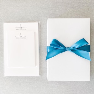May include: Two white notepads with a blue ribbon tied around them. The notepads have the text "ks" and "Kada Sweeten" printed on them.