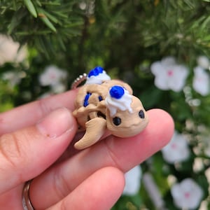 May include: A tan-colored turtle figurine with a dessert-like design. The turtle has a light brown shell and head, with blue and white accents. The figurine is held in a hand, with a blurred green and white floral background.