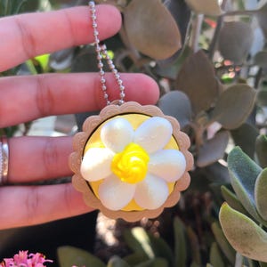 May include: A daisy-shaped keychain charm. The charm features a yellow center and white petals, set against a yellow background within a scalloped, tan border. A silver ball chain is attached for hanging.