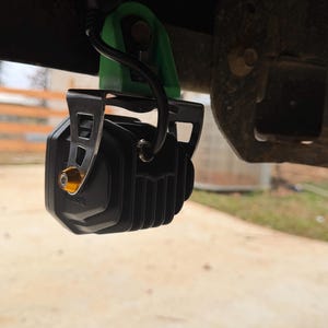 May include: A black LED light with a ribbed design, mounted with a green bracket and metal frame. The light has a gold-colored bolt and a black cable. The light is attached to a vehicle component.