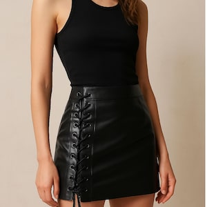 May include: Black faux leather mini skirt with a lace-up detail on the side. The skirt is paired with a black ribbed tank top. The skirt has a straight hemline and a high waist. The outfit is a classic and versatile style.