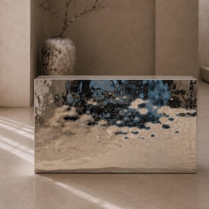 Könnte beinhalten: Ein rechteckiger Konsolentisch mit einer hochglanzpolierten, reflektierenden silbernen Metalloberfläche, die eine wellige, wasserartige Textur aufweist und verzerrte blaue und dunkelgraue Reflexionen auf der Vorderseite erzeugt.