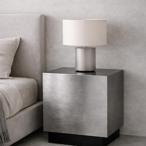 Può includere: Una scena di camera da letto moderna con un comodino in metallo spazzolato, una lampada cilindrica e un paralume bianco. Il comodino ha una base nera. È visibile anche un letto con testiera e biancheria da letto grigie.