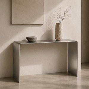 Könnte beinhalten: Ein Konsolentisch aus gebürstetem Metall mit glattem, geschwungenem Wasserfall-Design. Auf der Oberfläche stehen eine kleine, runde graue Schale und eine hohe, strukturierte beige Vase mit einer zarten Pflanze mit feinen Zweigen.