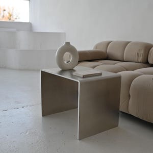 Könnte beinhalten: Ein modernes Wohnzimmer mit einem Beistelltisch aus gebürstetem Metall, einer weißen, kreisförmigen Vase und einem kleinen Buch. Im Hintergrund befindet sich ein beiges Ecksofa. Der Raum hat weiße Wände und Böden.