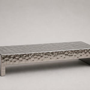 Pode incluir: Uma bandeja retangular de metal prateado com textura martelada. A bandeja tem um perfil baixo com um topo plano e duas pernas curtas. A superfície tem um padrão de covinhas, e o design geral é moderno e minimalista.