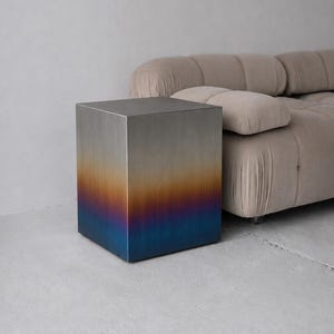 Peut inclure: Une table d'appoint carrée en métal avec une finition brossée. La surface présente un dégradé de couleurs vertical qui passe d'un argent métallique sur le dessus, à travers des tons orange et violet, jusqu'à un bleu profond à la base du cube.