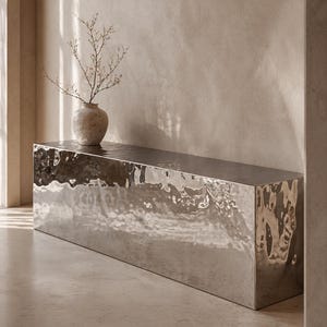 Pode incluir: Uma moderna consola prateada retangular com uma superfície de metal martelado texturizado. Um pequeno vaso texturizado com ramos secos em cima. A mesa está numa sala de tons neutros. A mesa tem cerca de 150 cm de comprimento.