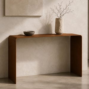 Könnte beinhalten: Ein brauner, U-förmiger Konsolentisch mit glattem, geschwungenem Design. Auf dem Tisch stehen eine kleine, dunkelgraue Schale und eine strukturierte, helle zylindrische Vase mit einem dünnen, kahlen Zweig.