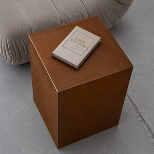 Peut inclure: Une table d'appoint carrée en métal de couleur rouille avec une finition mate lisse. Sur la surface supérieure repose un petit livre rectangulaire beige clair avec le texte LOUIS VUITTON imprimé en lettres dorées sur la couverture.