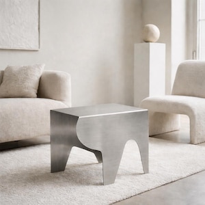 Może przedstawiać: Nowoczesny stolik boczny ze szczotkowanego metalu o unikalnym, zakrzywionym kształcie nóg. Kwadratowy stół wykonany jest ze stali nierdzewnej i stoi na białym dywanie. Nowoczesny mebel.