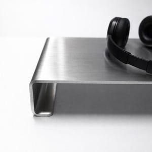 Könnte beinhalten: Schwarzer Over-Ear-Kopfhörer liegt auf einem Stand aus gebürstetem Metall. Der Ständer hat ein minimalistisches Design mit einer gebogenen Kante und einer rechteckigen Form. Der Kopfhörer befindet sich auf der rechten Seite.