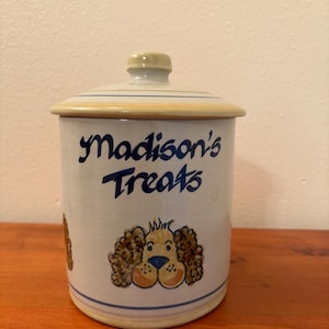 May include: A ceramic dog treat jar with a lid. The jar is white with blue accents and features the text "Madison's Treats" in blue script. A cartoon dog face is on the front. The lid is beige with a blue stripe.