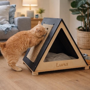 Puede incluir: Un gato jengibre se rasca en una casa para gatos de madera con techo negro y rascador de sisal. La casa para gatos tiene una cama dentro y el nombre "Luna" grabado en el lateral. Dos cuencos de acero inoxidable están unidos a la base.