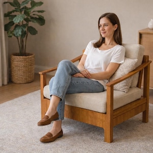 May include: A wooden armchair with a cream-colored cushion and a light-colored wooden frame. The chair is in a well-lit room with a rug and a potted plant. The person is wearing a white t-shirt, blue jeans, and brown loafers.