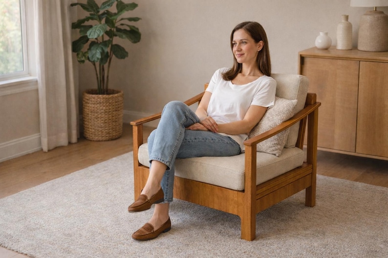 May include: A wooden armchair with a cream-colored cushion and a light-colored wooden frame. The chair is in a well-lit room with a rug and a potted plant. The person is wearing a white t-shirt, blue jeans, and brown loafers.