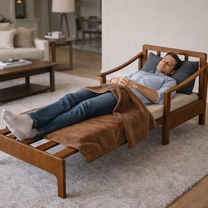 May include: Wooden chaise lounge with a brown blanket. The lounge has a light-colored cushion and two gray pillows. The frame is made of wood. The lounge is in a living room with a rug and a sofa.