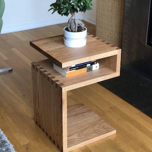 May include: A wooden side table with a unique, open design. The table features a top surface with a small bonsai tree in a white pot. The table has a lower shelf with books. The wood has a natural grain and a warm, light brown color.