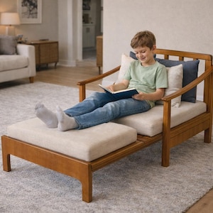 May include: A wooden chaise lounge with a light beige cushion and a matching ottoman. The chair has a light brown wooden frame and is placed on a gray rug. A person is sitting on the chair reading a book.