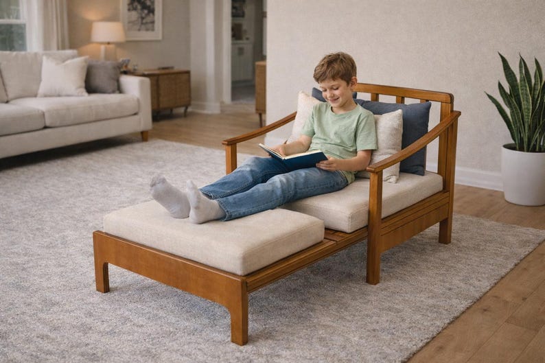 May include: A wooden chaise lounge with a light beige cushion and a matching ottoman. The chair has a light brown wooden frame and is placed on a gray rug. A person is sitting on the chair reading a book.