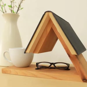 May include: A wooden book stand with a book forming a roof shape, holding a pair of eyeglasses. A white mug and a vase with greenery are also on the wooden surface. The stand is made of light-coloured wood.