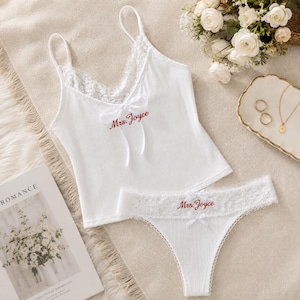 Könnte beinhalten: Ein weißes, geripptes Camisole- und String-Set mit zarter weißer Spitzenborte am Ausschnitt und am Bund. Beide Teile sind mit einer kleinen weißen Schleife und dem in Rot gestickten Schriftzug Mrs. Joyce auf der Vorderseite versehen.