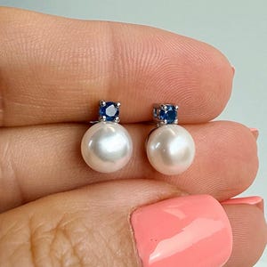 Op de afbeelding: Een paar oorknopjes met ronde witte parels bekroond door kleine, gefacetteerde blauwe edelstenen gezet in zilverkleurig metaal. Elke oorbel bestaat uit een parel met een blauw steentje dat direct boven het oppervlak van de parel is geplaatst.