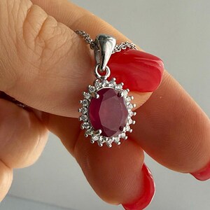 May include: A silver-toned pendant necklace featuring an oval-shaped deep red gemstone at the center, surrounded by a halo of small, clear, sparkling stones. The pendant is attached to a delicate, textured silver-toned chain link necklace.