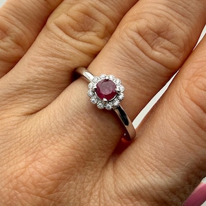 Puede incluir: Un anillo de plata con una piedra preciosa central redonda de color rojo oscuro, rodeada por un halo de piedras preciosas más pequeñas y claras. El anillo está sobre un fondo claro.
