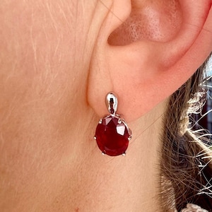 Può includere: Un orecchino singolo caratterizzato da una gemma rosso scuro di forma ovale sfaccettata, montata su una struttura in metallo color argento lucido. L'orecchino è fissato a un piccolo gancio liscio color argento che si collega al lobo dell'orecchio.