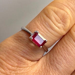 Könnte beinhalten: Ein silberner Ring mit einem rechteckigen, tiefroten Edelstein. Der Ring hat ein schlichtes Design mit einem schmalen Band und einem auffälligen Stein. Die Farbe des Edelsteins ist ein sattes, leuchtendes Rot.