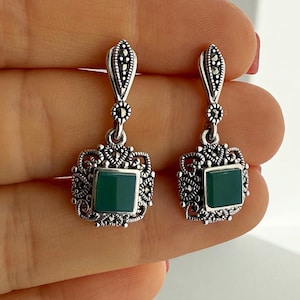 Puede incluir: Un par de pendientes colgantes con piedras verdes cuadradas engastadas en metal plateado. Cada pendiente tiene un marco ornamentado de estilo filigrana con pequeñas piedras oscuras facetadas alrededor de la piedra central.