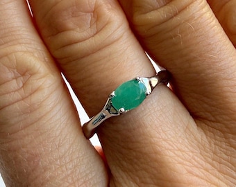 May include: A silver ring featuring a vibrant green oval-cut gemstone. The ring's band is slender, and the gemstone is set in a classic, minimalist style. The ring is shown on a hand with manicured nails.