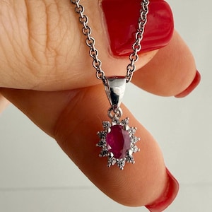 Peut inclure: Un collier argenté doté d'un pendentif en pierre précieuse rouge foncé de forme ovale, entouré d'un halo de petites pierres claires et scintillantes. Le pendentif est fixé à une fine chaîne argentée aux maillons circulaires.
