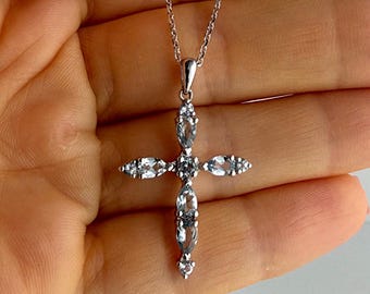 Puede incluir: Un collar de tono plateado con un colgante de cruz adornado con múltiples piedras preciosas de color azul claro en talla marquesa dispuestas en un patrón vertical y horizontal, suspendido de una delicada cadena de metal plateado.