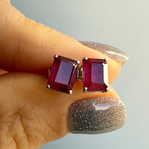 Puede incluir: Un par de pendientes de botón con piedras preciosas rectangulares de color rojo intenso engastadas en plata. Las gemas tienen un color rico y saturado y están montadas sobre un engaste sencillo y elegante. Los pendientes se muestran sobre un fondo azul claro.