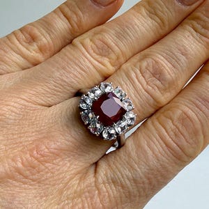 Könnte beinhalten: Ein silberner Ring mit einem großen, quadratisch geschliffenen, tiefroten Edelstein, umgeben von kleineren, klaren Steinen. Das Design des Rings ist klassisch und elegant, mit Fokus auf dem zentralen rubinähnlichen Stein.