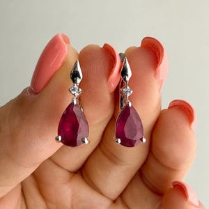 Pode incluir: Um par de brincos pendentes de prata com pedras preciosas vermelho-escuras em forma de pera suspensas por um pequeno detalhe de pedra clara. Os brincos têm um acabamento metálico polido e um fecho de dobradiça seguro.