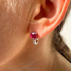 May include: A close-up of a silver earring with a square, deep red gemstone. The earring has a small hoop design with a row of tiny, clear stones. The earring is worn on a person's ear.