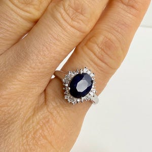 Hand holding a sterling silver ring with a deep blue oval sapphire in a ballerina halo, stones radiating outward in a crown pattern