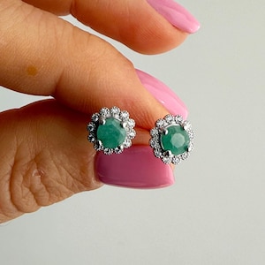 Puede incluir: Un par de pendientes de botón con una piedra preciosa central redonda de color verde esmeralda rodeada por un halo de piedras preciosas claras más pequeñas. Los pendientes están engarzados en un metal plateado y se muestran sobre un fondo claro.