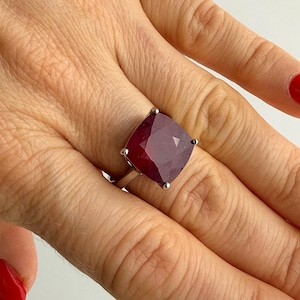 May include: A silver ring featuring a large, square-cut, deep red gemstone with faceted surfaces. The stone is set in a four-prong metal setting on a simple, polished silver band, showcasing a bold and prominent design on the finger.