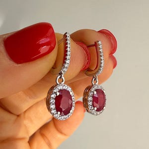 May include: A pair of silver drop earrings. Each earring has a large, oval, deep red gemstone surrounded by a halo of small, clear gemstones. The earrings are held up by a lever back closure.
