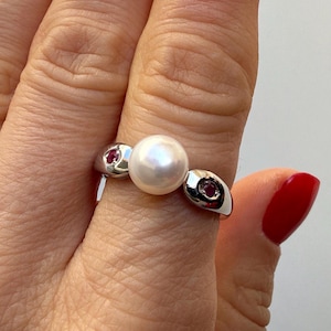 Può includere: Un anello d'argento con una grande perla bianca rotonda come pietra centrale. Due montature ovali più piccole su ciascun lato contengono ciascuna una piccola gemma rossa. L'anello è mostrato su un dito.