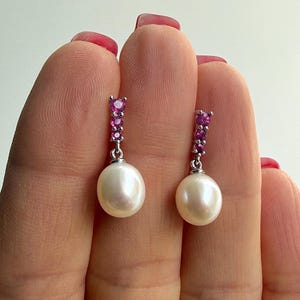 Könnte beinhalten: Ein Paar elegante Ohrringe mit einer großen, cremeweißen Perle, die unter einer vertikalen Reihe kleiner, leuchtend rosa Edelsteine hängt. Die Ohrringe sind in einem silberfarbenen Metall gefasst und wirken klassisch und edel.