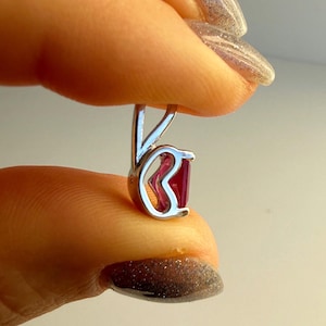 Könnte beinhalten: Ein silberner Ohrring mit einem leuchtend roten Edelstein. Der Ohrring hat ein einzigartiges Design mit einer geschwungenen Form, die den Edelstein hält. Der Edelstein hat eine tiefrote Farbe und verleiht dem Schmuckstück einen Farbtupfer. Der Ohrring wird zwischen Fingern gehalten.